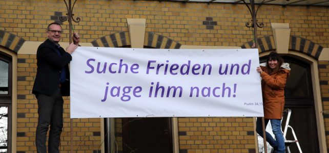 Bischof Thomas Adomeit (li.) und Birgit Carmona Scheider vom Bischofsbüro befestigen das Banner mit den Psalmworten „Suche Frieden und jage ihm nach!“ (Psalm 34,15) am Gebäude des Oberkirchenrates im Oldenburger Philosophenweg. Foto: ELKiO/D.-M. Grötzsch